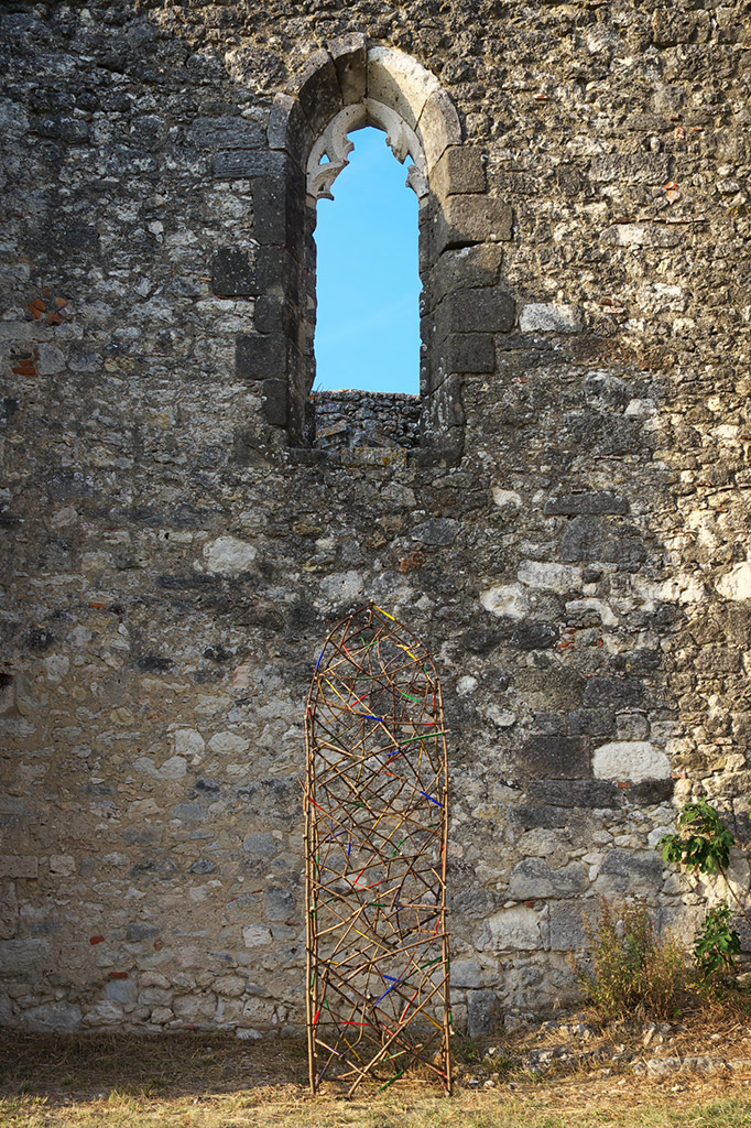Structure tressée en bois installée contre un mur en pierre d’un bâtiment ancien, à côté d’un arbre feuillu.