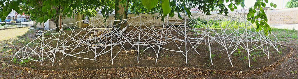 Sous un arbre, sur un sol en terre, se trouve une sculpture de plusieurs mètres de long en bois blanc de l'artiste Anne Mangeot.