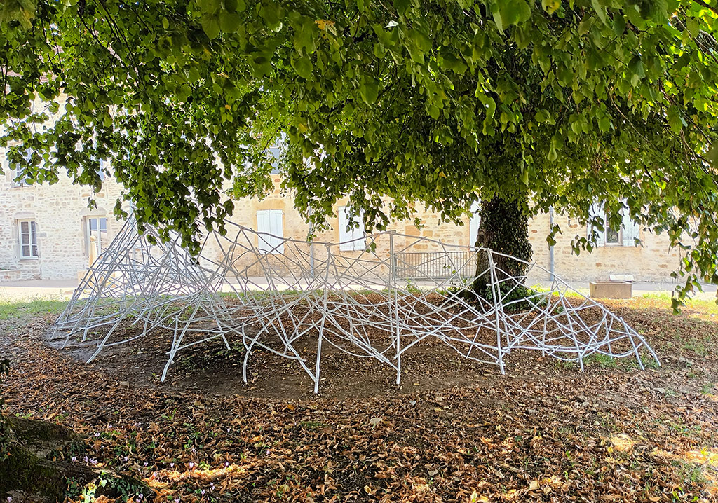 Dans un jardin devant un ancien hôpital, une sculpture en bois de châtaignier est installée sous des arbres.