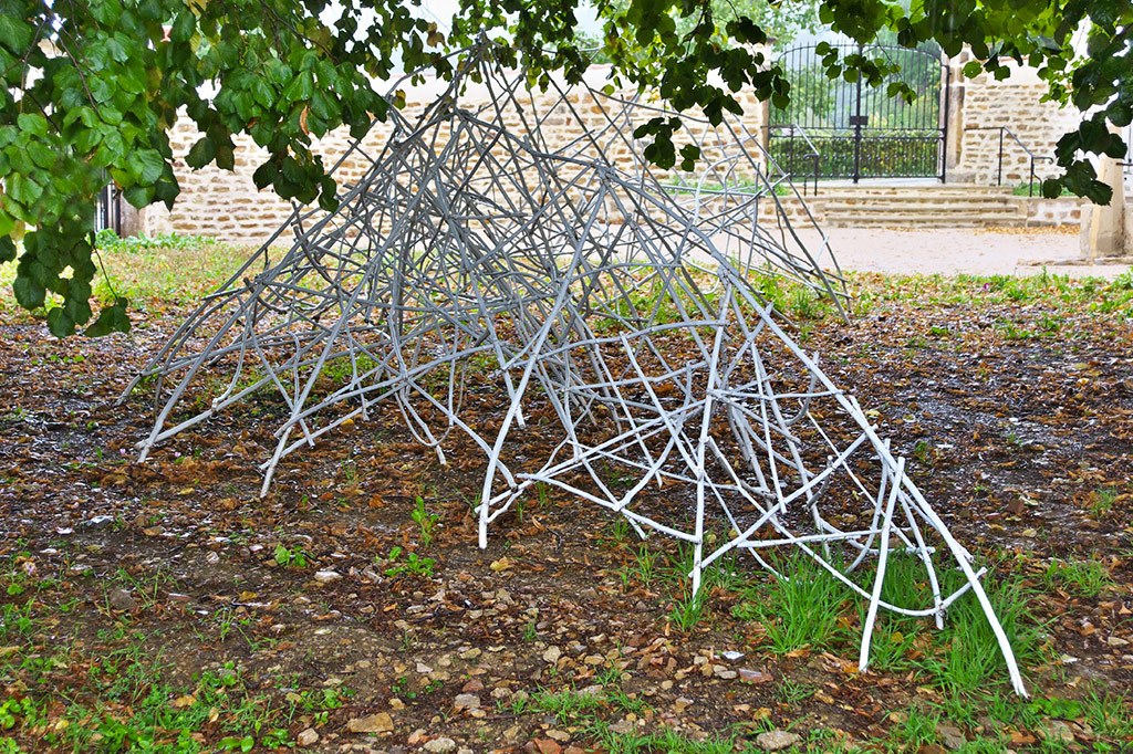 Dans un jardin devant un ancien mur en pierre se trouve une sculpture en bois de châtaignier, de profil, posée sur le sol et sous des branches d'arbres.