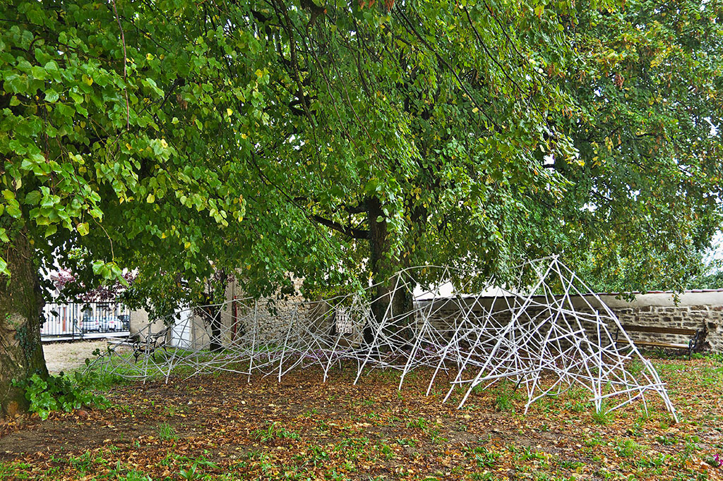 Au loin, devant un mur de pierre et sous de grands arbres se trouve une grande sculpture faite de bois de châtaignier.