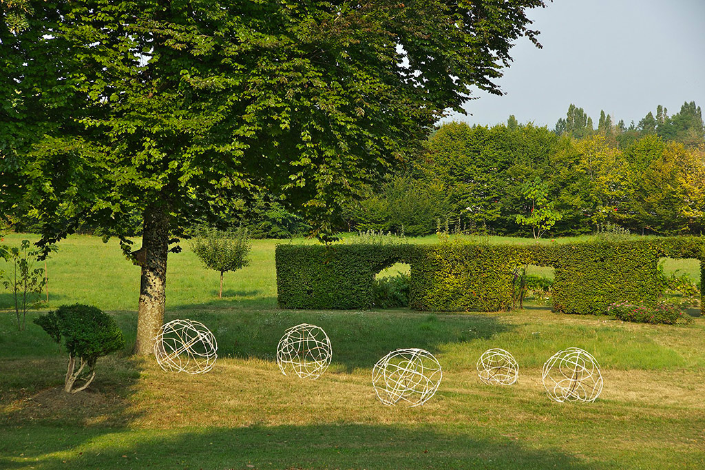 Installation de l'artiste Anne Mangeot, cinq structures de boules schématiques faites en branchages sont installées, au loin, sous un grand arbre, dans un parc. 