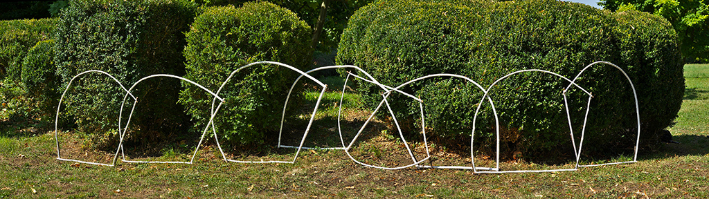 Installation de l'artiste Anne Mangeot, aux formes géométriques évidées, faite de branchage blancs, apposé contre des buissons en extérieur.
